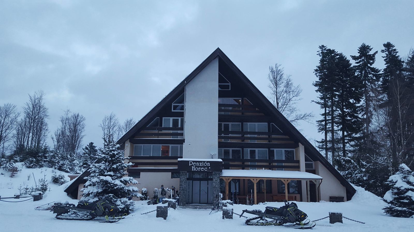 Hotel Horec v zime za súmraku: strmá strecha v tvare A, rozsvietené okná, pred vchodom snežné skútre.