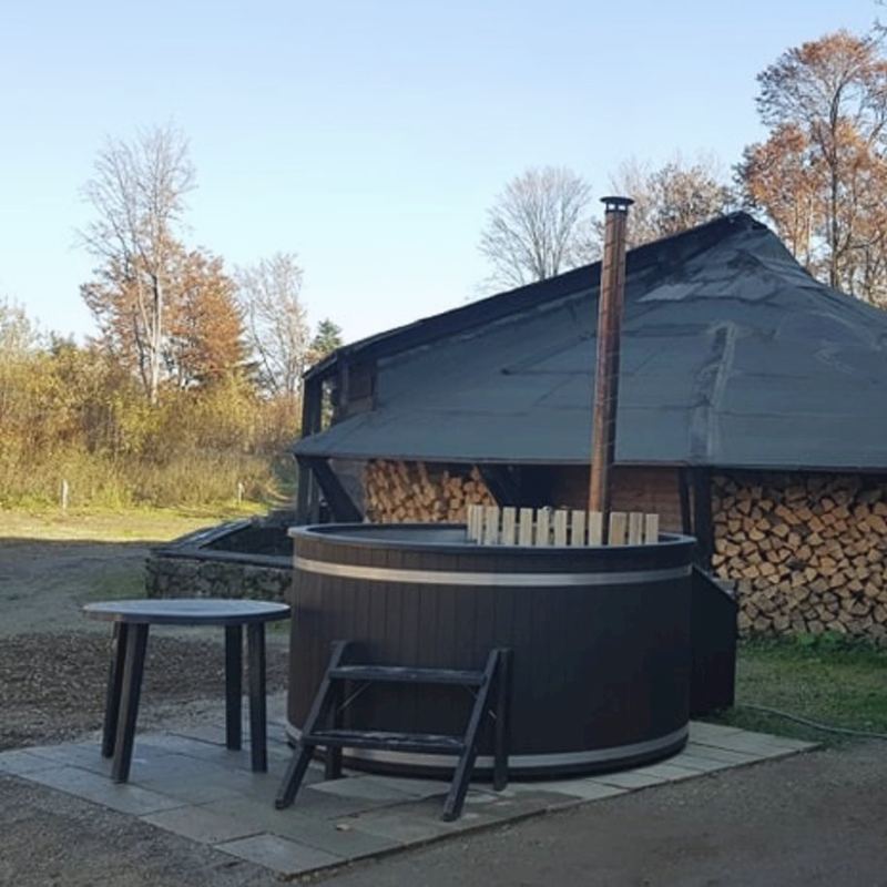 Drevený kúpací sud s komínom pod prístreškom, vedľa naukladané drevo a jesenné stromy.